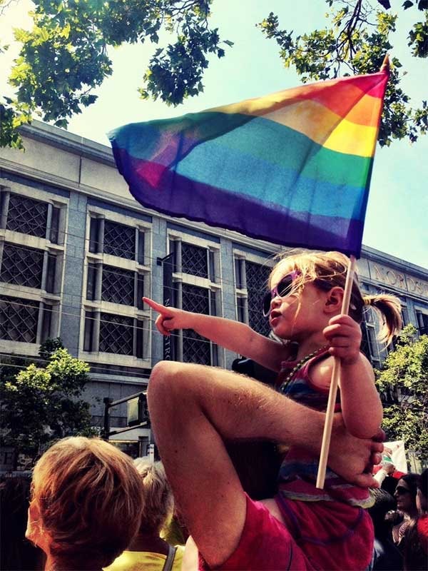 child on parent's shoulder holding rainbow flag